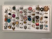 Top-down view of over 50 assorted costume rings displayed on white board, showing wide variety of styles and colors.