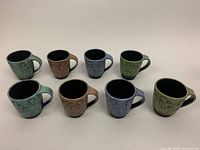 Eight assorted colored pottery mugs arranged, displaying handles and exterior etchings.