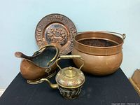 Four antique metal items on a black cloth, including a decorated brass Moroccan tea kettle, copper scuttle, cauldron, and decorative wall plate