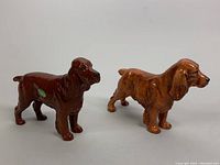 Two porcelain Cocker Spaniel dog figurines, one dark brown, one lighter brown, displayed side by side on a neutral background