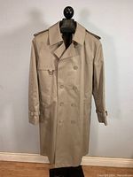 Full front view of the beige trenchcoat on a mannequin against a white wall and wooden floor.