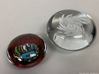 Two blown glass paperweights on a white surface, one clear with white swirl and one red with multi-color center