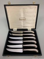 Photo showing original black presentation box lined with satin and velvet, containing six cutting knives with silver handles arranged in slots.