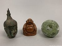 Front view of bronze Thai Buddha head, wooden happy Buddha sculpture, and jade interlocking puzzle ball.