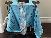 Front view of three black metal spiral candle stands together with a blue cloth backdrop, one stand covered in plastic wrap.