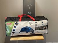 Side view of Coleman Arrowhead 8-person tent in black carrying bag with red handles, showing label with tent image and features.
