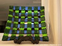 Front view of square glass plate with green and blue basket weave pattern displayed on ornate dark metal stand.