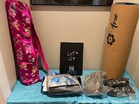 Fitness lot items shown against a beige wall on a blue mat including floral pink yoga mat bag, cork yoga mat with Aire branding, AbTronics kit, and two fitness balls.