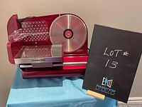 Front and side views of the red Kalorik electric food slicer showing the large round blade, food carriage, thickness adjustment dial, and safety pusher.