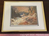 Framed artwork showing a landscape print of river falls surrounded by autumn trees and rocks in a light frame.