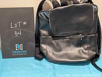 Black knapsack purse with front flap and zippered pocket shown front view with contest sign.