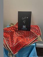 Two decorative pillow covers draped over a table, showing red fabric with gold circular patterns and thick gold braided edges with tassels.