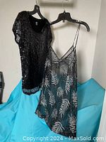 Front view of Joe Fresh black sequin top and long black transparent sparkle camisole hanging on hangers against white wall and turquoise background.