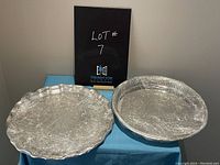 Two round silver plate serving trays wrapped in plastic on a blue surface, photo shows overall lot contents