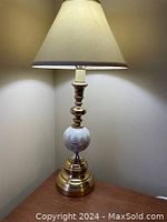 Front view of a brass-colored table lamp with a beige fabric shade on, showing the detailed base and lampshade condition.