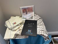 Lot with a white lace tablecloth, beige embroidered tablecloth, and floral vinyl tablecloth included along with lot identification sheet and packaging.