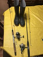 Photo of two fishing rods laid parallel on a yellow surface, with their respective spinning reels attached and a pair of black rubber Wellington boots standing upright.