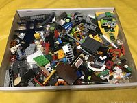 Top view of a cardboard box filled with assorted LEGO pieces including bricks, small plates, windows, and wheels in various colors.