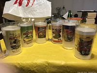 Six assorted collectible plastic mugs from Autofest show displayed on yellow cloth, showing full designs including cars and event text.