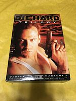 Front cover of Die Hard Trilogy DVD box set showing Bruce Willis holding a gun, shirtless in tank top, with night sky background.