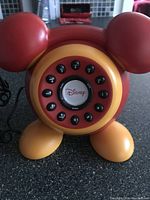 Front close-up of Disney rotary dial phone showing red ears, yellow feet and central rotary dial with Disney logo