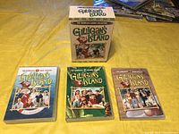Overview of the Gilligan's Island DVD series collection items on yellow fabric background, showing the box and three DVD cases for different seasons.