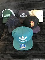 Lot of six caps displayed on black fabric background showing front designs and logos
