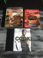 Three cookbooks laid on floor: Jamie Oliver's 'Cook with Jamie' in the foreground, 'The Complete Family Cookbook' and 'Delicious & Dependable Slow Cooker Recipes' in the background.