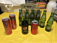 Photo showing six green glass Canada Dry Millennium bottles with three vintage soda cans (two Coca-Cola and one Pepsi) on yellow surface.