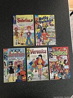 Five Archie comic books arranged on a dark surface showing covers of Sabrina, Betty and Veronica, Archie, Veronica, and Jughead comics.