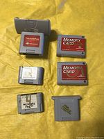 Six Nintendo 64 memory and accessory cartridges laid out on yellow background showing labels and handwritten markings, including two Memory Cards and one TremorPak.
