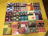 All 15 paperback novels laid out on yellow surface showing the colorful covers of each book with character photos and graphic patterns.