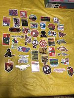 Photo showing full collection of about 44 Woodstock festival-themed stickers arranged on yellow fabric background. Stickers vary in size from 2 to 3 inches with colorful designs including guitars, peace signs, people, vans, and festival logos.