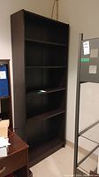 Full view of a dark brown manufactured wood bookcase with five shelves, placed against a beige wall next to an office desk and filing cabinet.