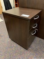 Full view of the laminated filing cabinet showing dark wood finish, three silver metal handles, plastic casters under the cabinet, and minor top surface mark.