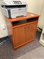 Closed view of reddish-brown laminated particle board cabinet with flat top and two-door lower storage, open shelf above doors, placed on carpeted floor with printer on top.