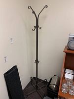 Front view of tall metal coat rack stand with four splayed legs and multiple hooks, placed against a wall near a speaker and waste bin.