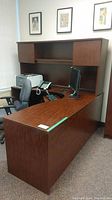 Full view of L-shaped dark wood grain manufactured wood office desk with hutch and storage compartments, including a chair, monitor, keyboard, and phone (contents not included).