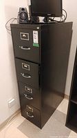 Full view of black metal filing cabinet with four drawers, locking mechanism on top drawer with keys attached, and hanging file folders inside. Cabinet is positioned next to a wall with a monitor and speakers placed on top.