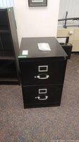 Front view of black metal 2-drawer filing cabinet showing scratched surface and drilled lock on top drawer.
