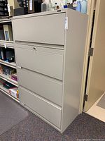 Full view of a beige metal filing cabinet with four drawers, closed and locked, placed beside shelving unit.