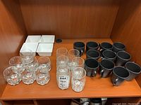 Shelf with 7 whiskey glasses, 6 highball glasses, 11 black ceramic mugs, and 4 white ceramic dip bowls