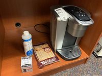 Photo showing Keurig Elite K90 coffee maker with descaling solution bottle, Keurig MY K-CUP reusable coffee filter box, and bamboo coffee stir sticks package on wooden shelf.
