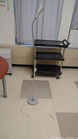 Full view of metal floor lamp standing on linoleum floor with rolling cart in background.