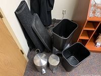 Photo showing a black textured floor mat rolled up next to a black plastic shoe tray, two black plastic waste bins, and two metal waste bins with foot pedals in front.