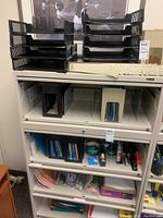 Multiple office supplies including stacked black plastic organizers on top shelves, paper filing guides with numbered tabs in front, shelves below holding binders, paper clips, staplers, and other supplies.