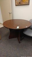 Photo showing the round manufactured wood office table with medium brown finish and a wide, curved base forming an X shape.