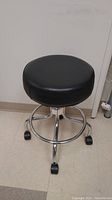 Front view of black vinyl cushioned rolling stool with chrome metal frame and caster wheels on a tiled floor.
