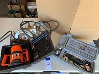 Black & Decker router in orange and black hard plastic case, Skil circular saw with beige metal body and circular blade on table with cardboard background.