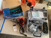 Overview of all items in the lot on a table: Dremel rotary tool kit in case, red handheld heat tool with cord, blue Makita circular saw box, and orange extension cord reel.